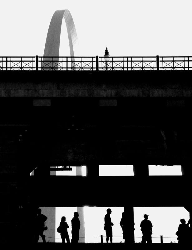 Black and white of a bridge in front of the St. Louis arch, people in dark relief waiting for a train.