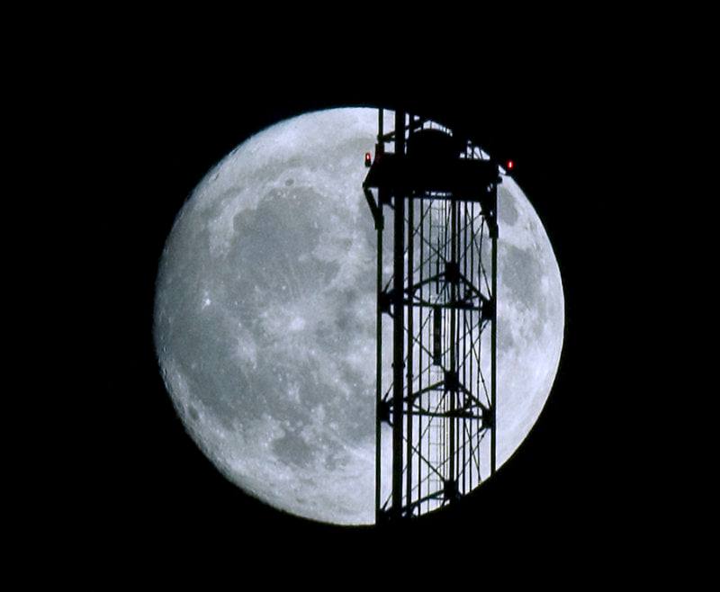 Full moon with tower in front with two red lights, one just at the edge of the moon. The tower partially obscures the moon, but with an open lattice work for the moon to shine through.