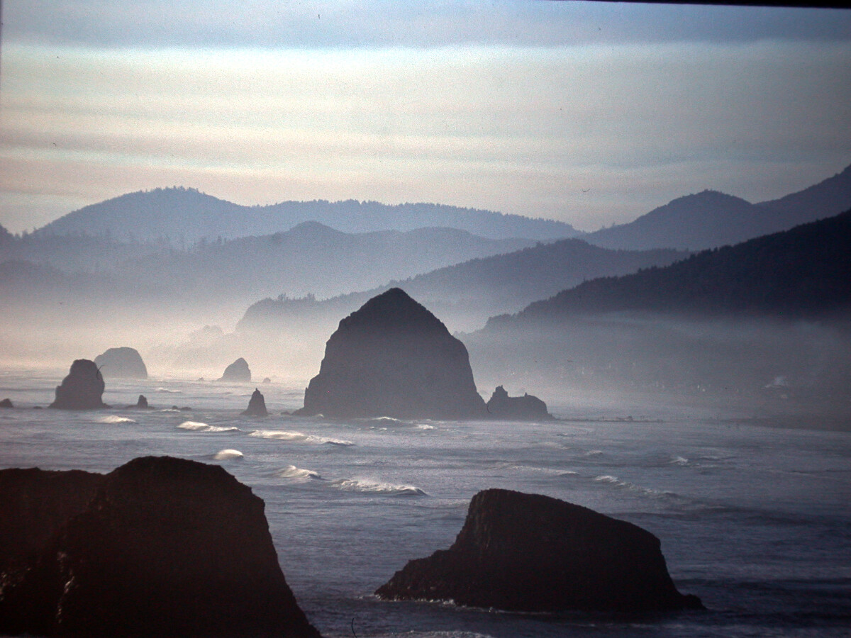 Hazy, fog laced series of low hills and large rocky outcroppings in ocean, including Haystack rock at Canon Beach but seen from a cliff further down from the beach. Gentle rolling ocean waves among the rocks.