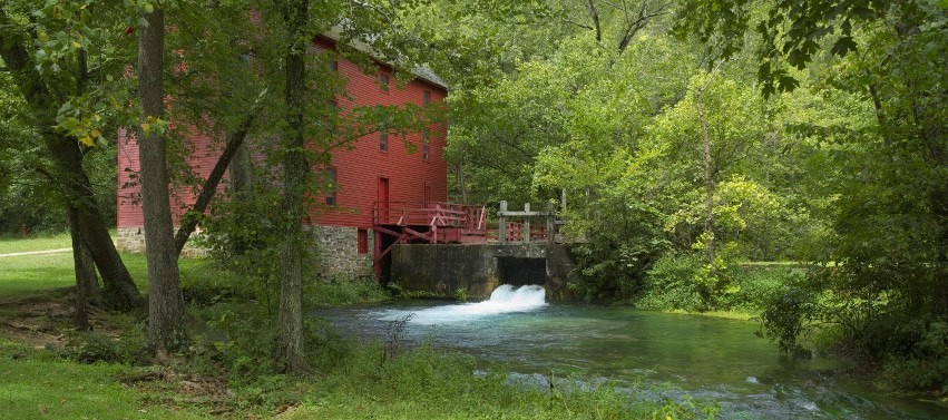 An old red mill with water coming through the sluice way in the midst of green trees. Afternoon shade, and the water is clear and slightly turquoise in color. Very peaceful.