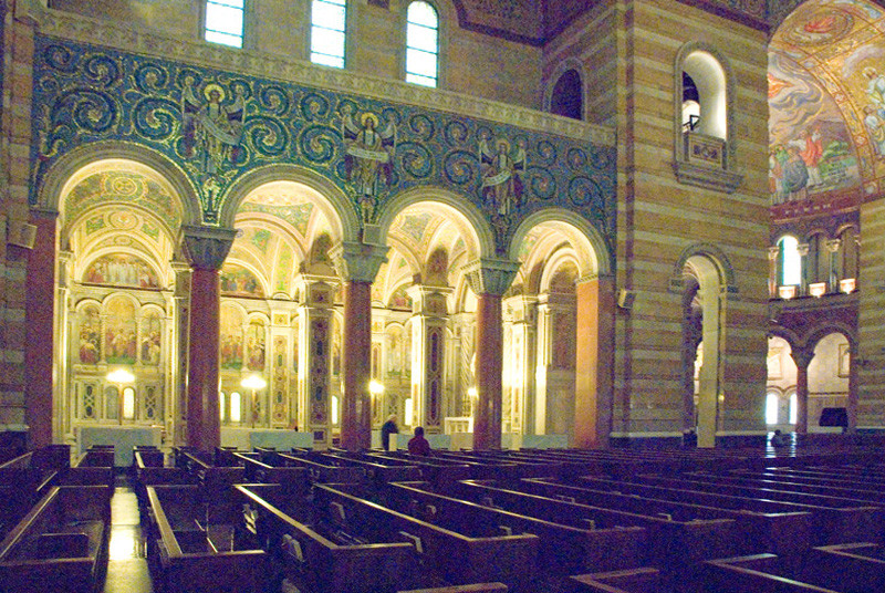 Inside a dark, indirectly lit cathedral with mosaic walls, wooden pews, granite columns, and a few people quietly talking in the back