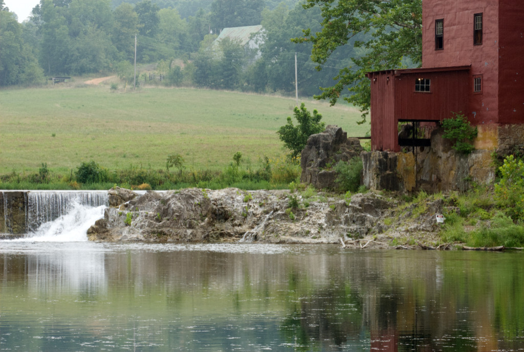 Old time mill, in red, with water fall next to it, and farm off in the distance with rolling hill tilted towards mill. Smooth retaining pond after waterfall.