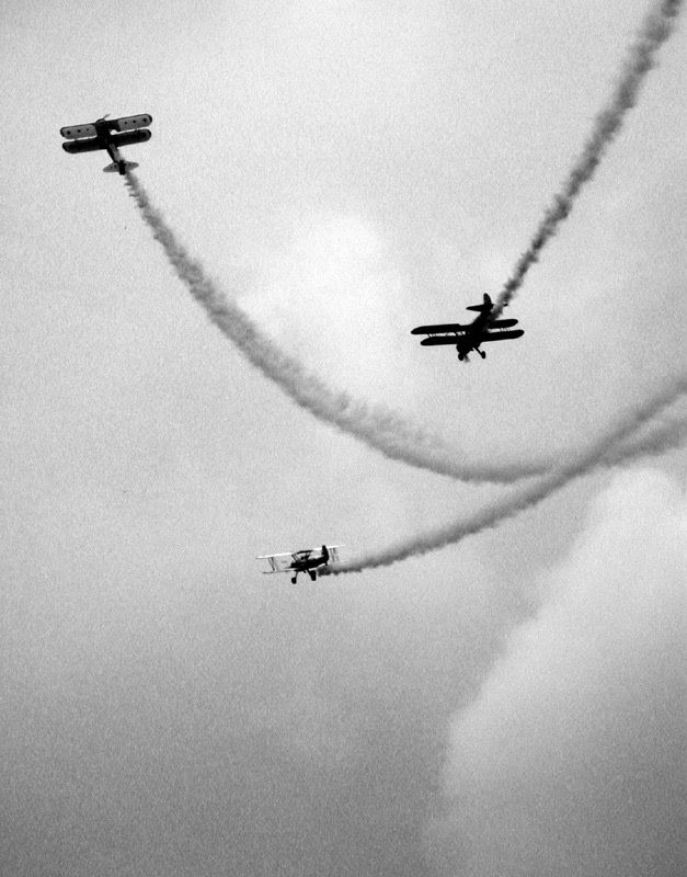 Three vintage biplanes, doing air stunts against a cloudy sky. All three planes are trailing smoke. Black and white photo.