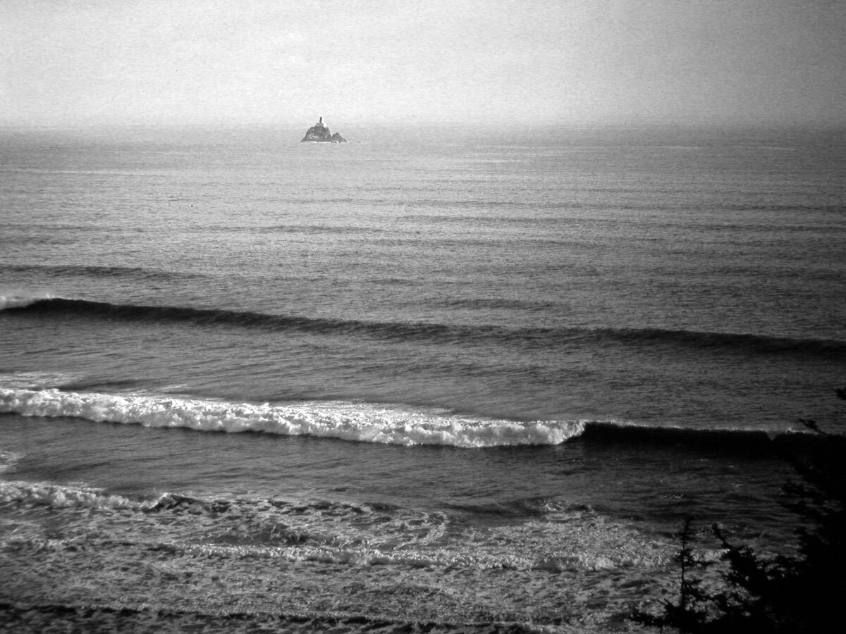 Black and white photo of distant lighthouse on small outcrop of rock, with gentle waves crashing against shore.