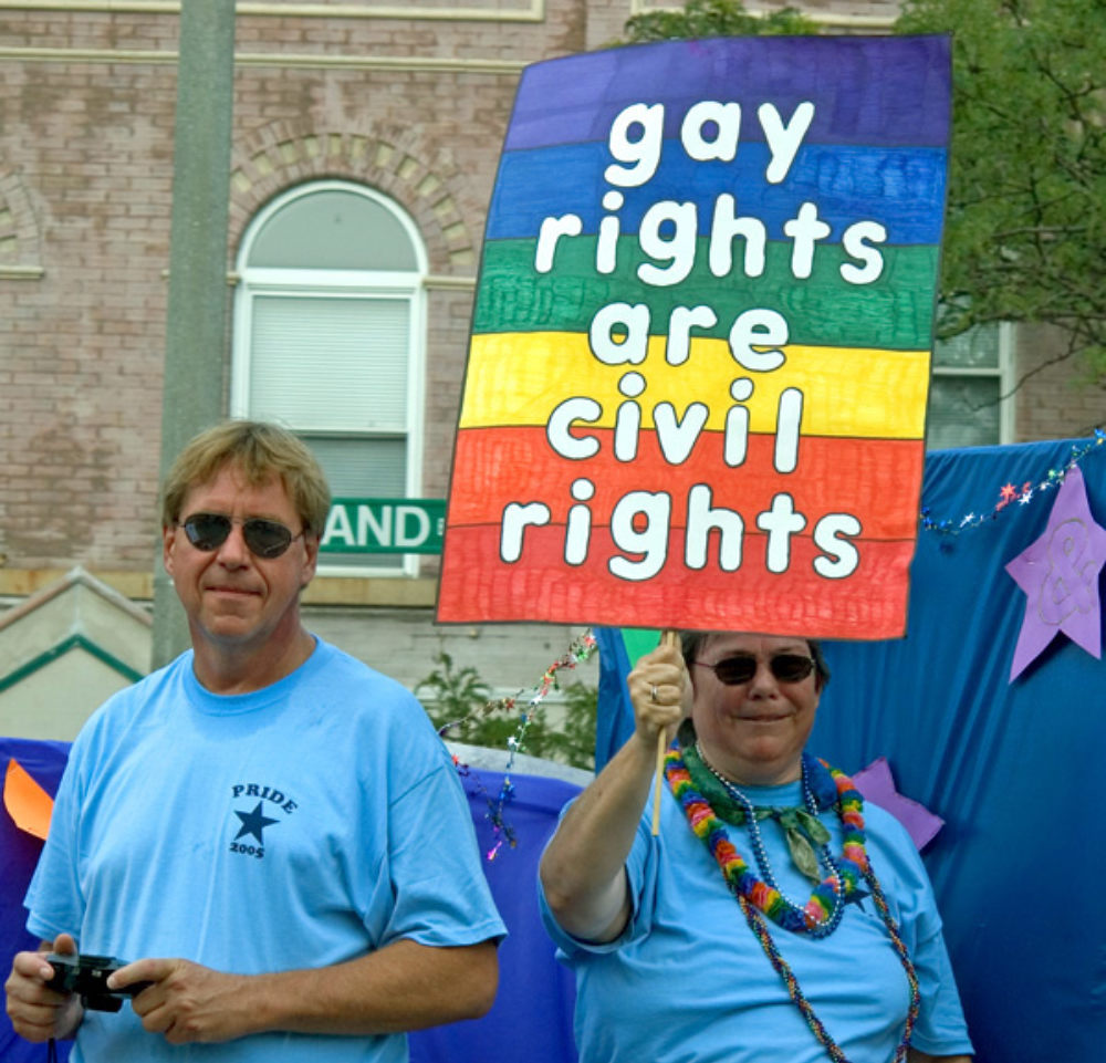 A man and a woman, both wearing bright blue shirts, looking at the camera and holding up a sign. The sign is painted in brilliant rainbow colors with the words, "gay rights are civil rights."