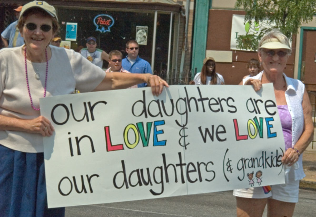 In Pride parade, two women marchers smiling at camera holding a hand made sign that reads "Our daughters are in LOVE and we LOVE our daughters (and our grandkids, too)" Both LOVES are in rainbow colors