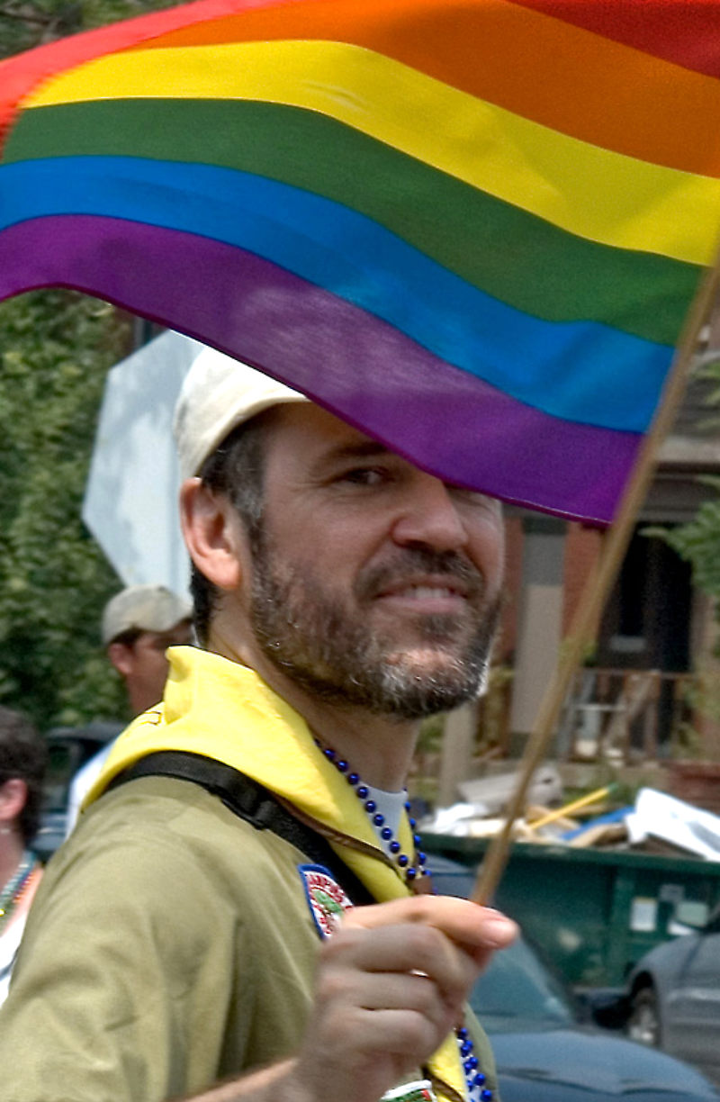 Man with beard dressed in Boy Scouts scoutmaster uniform, looking at camera, smiling, holding a pride flag that partially obscures his face.