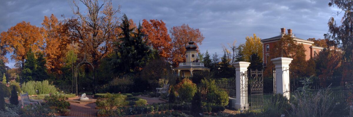 Panorama of a formal garden, showing fall colors, gate pillars and statue highlighted by late afternoon sunshine.