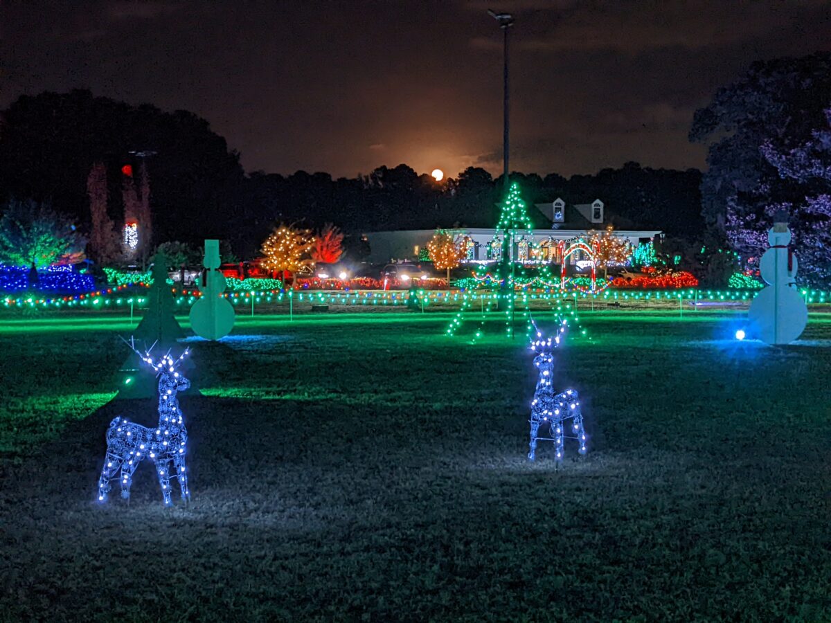 Christmas statues and lights in an open field with a full moon peeking out over trees in the background